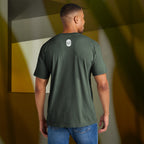 Man wearing a dark green t-shirt with an Eternal Optimist Golf Club logo on the back, standing against a geometric-patterned wall.