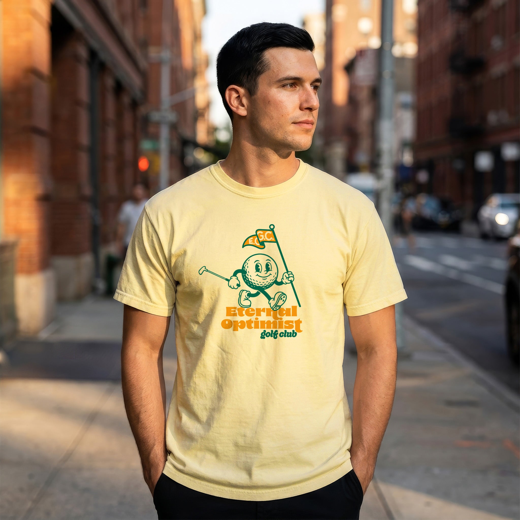 Man wearing a yellow t-shirt with a 1950s-inspired golf ball cartoon that has legs and arms. The cartoon golf ball is carrying a putter and a flagstick that has "E.O.G.C." on the flag. Below him are the words 'Eternal Optimist Golf Club'.