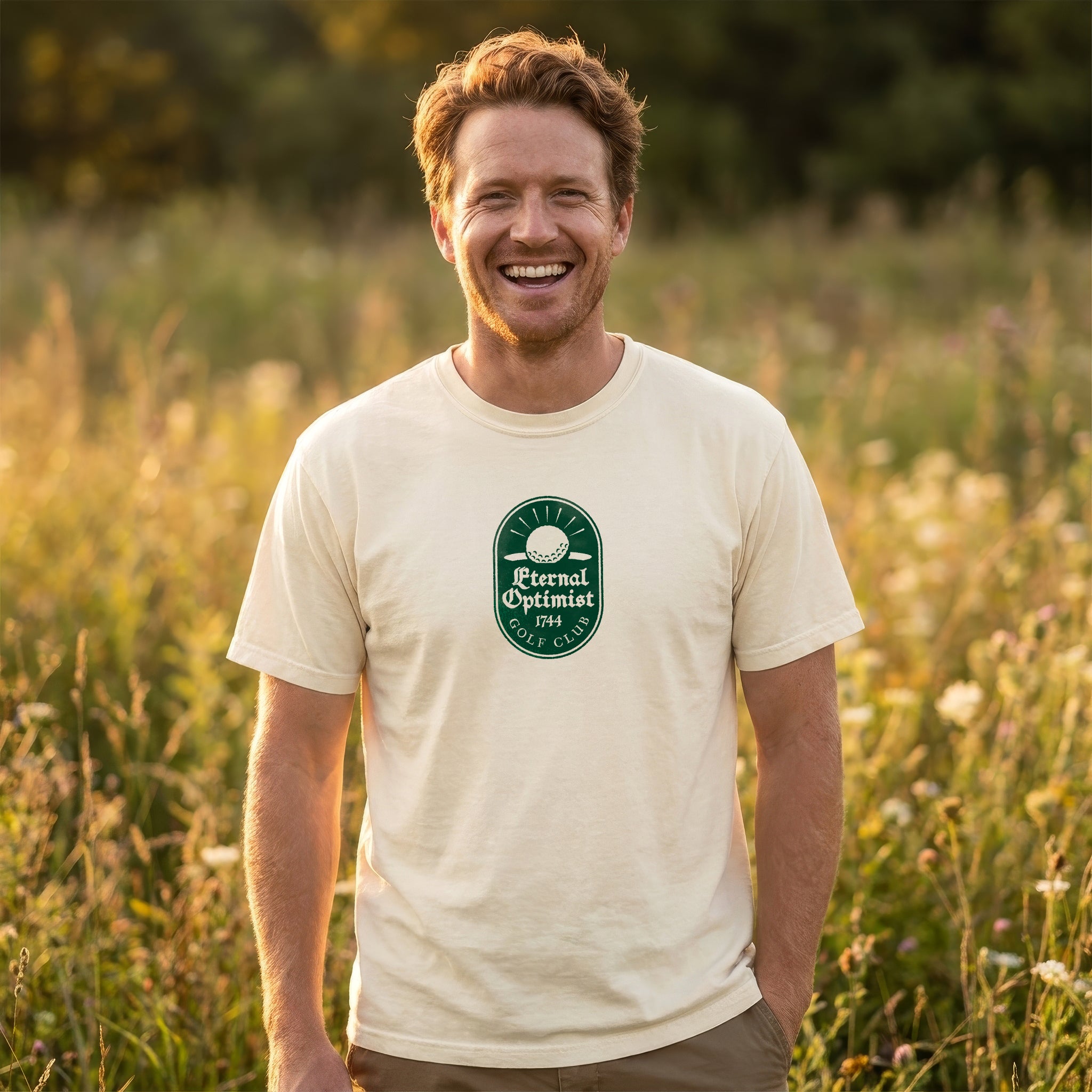 Red headed man, standing in a field of grass and wildflowers, wearing an Ivory t-shirt with a green Eternal Optimist Golf Club logo. 