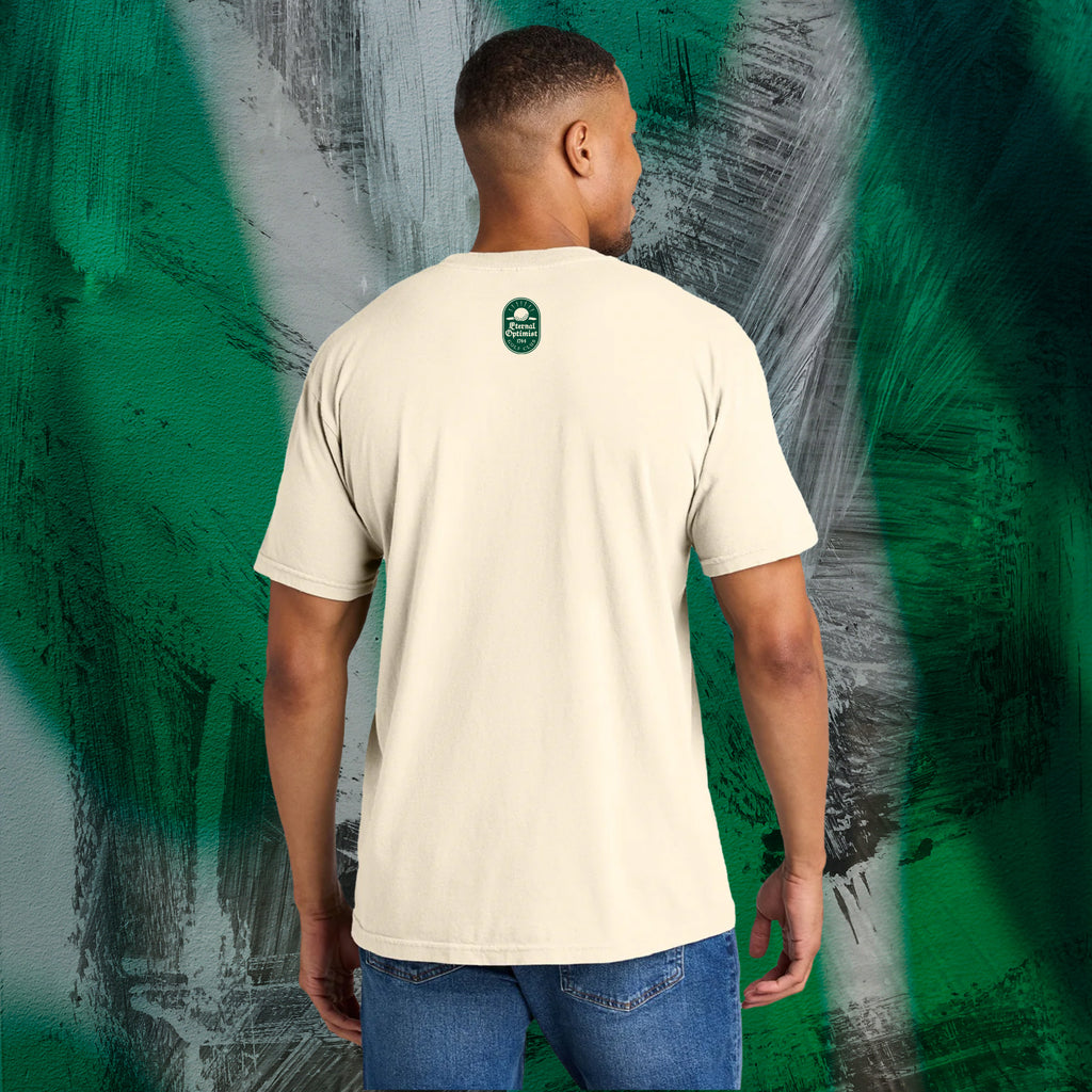 Man wearing an ivory t-shirt with a green Eternal Optimist Golf Club logo on the back, standing against an abstract-patterned wall.