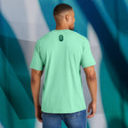 Man wearing a light green t-shirt with a green Eternal Optimist Golf Club logo on the back neck, standing against an abstract-patterned wall.
