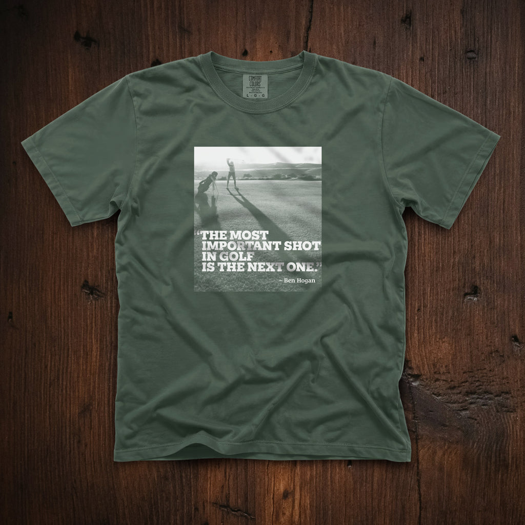 Dark green t-shirt that has an image of a golfer with a quote from Ben Hogan: "The most important shot in golf is the next one."