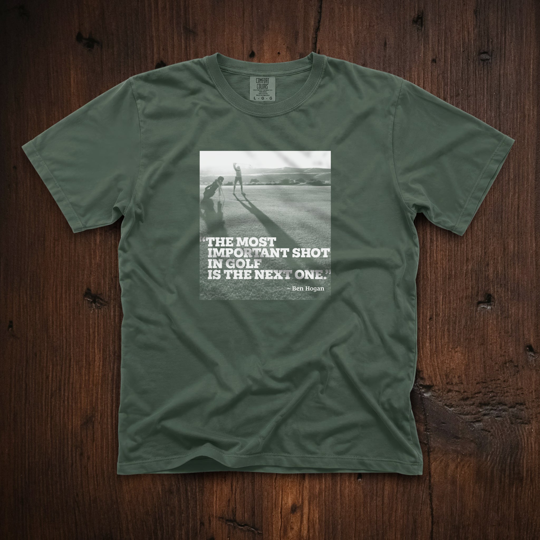 Dark green t-shirt that has an image of a golfer with a quote from Ben Hogan: "The most important shot in golf is the next one."