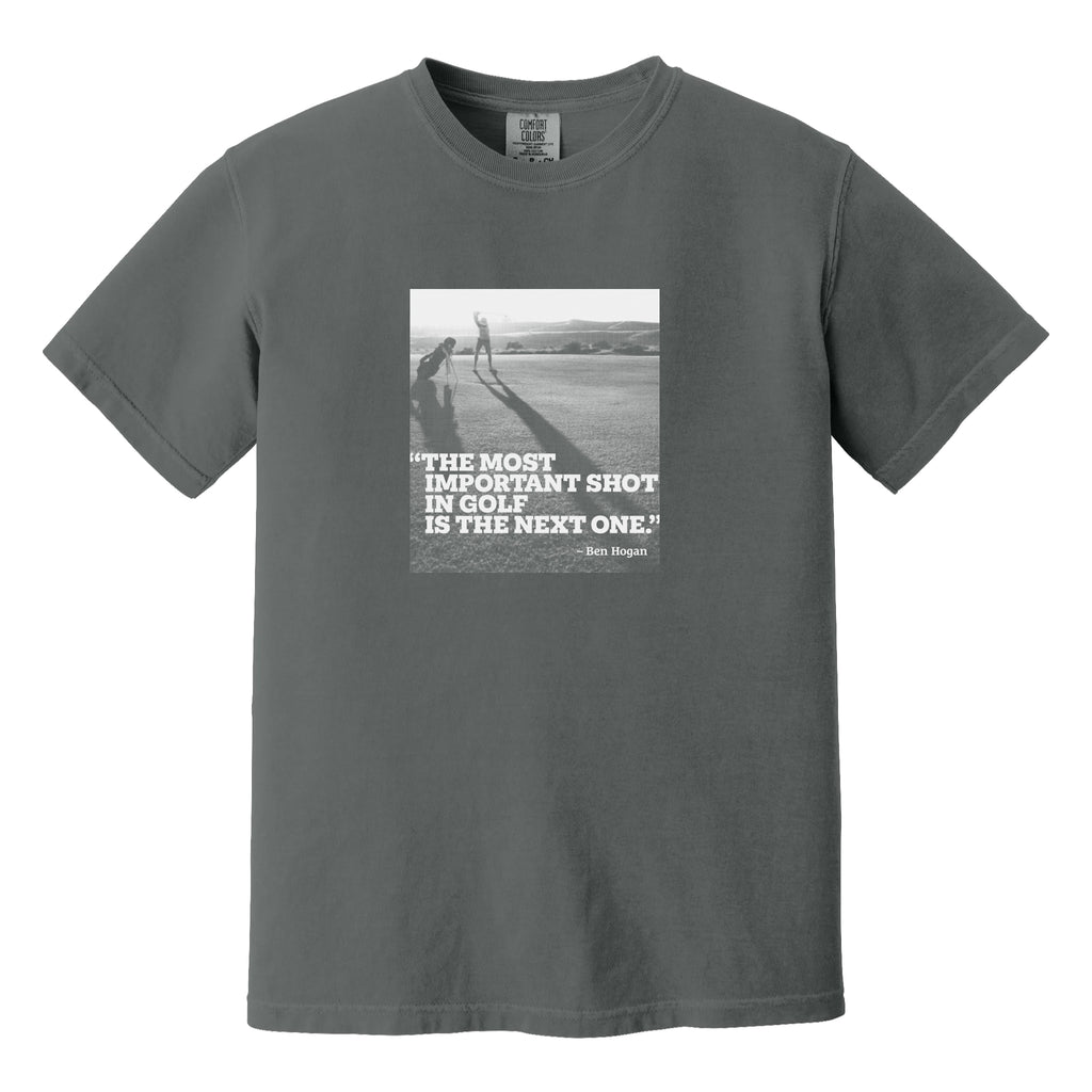 Dark gray t-shirt that has an image of a golfer with a quote from Ben Hogan: "The most important shot in golf is the next one."