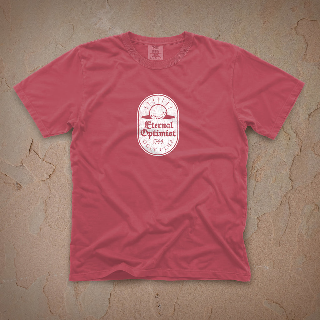 Dark pink t-shirt with a white Eternal Optimist Golf Club logo on a beige textured background.