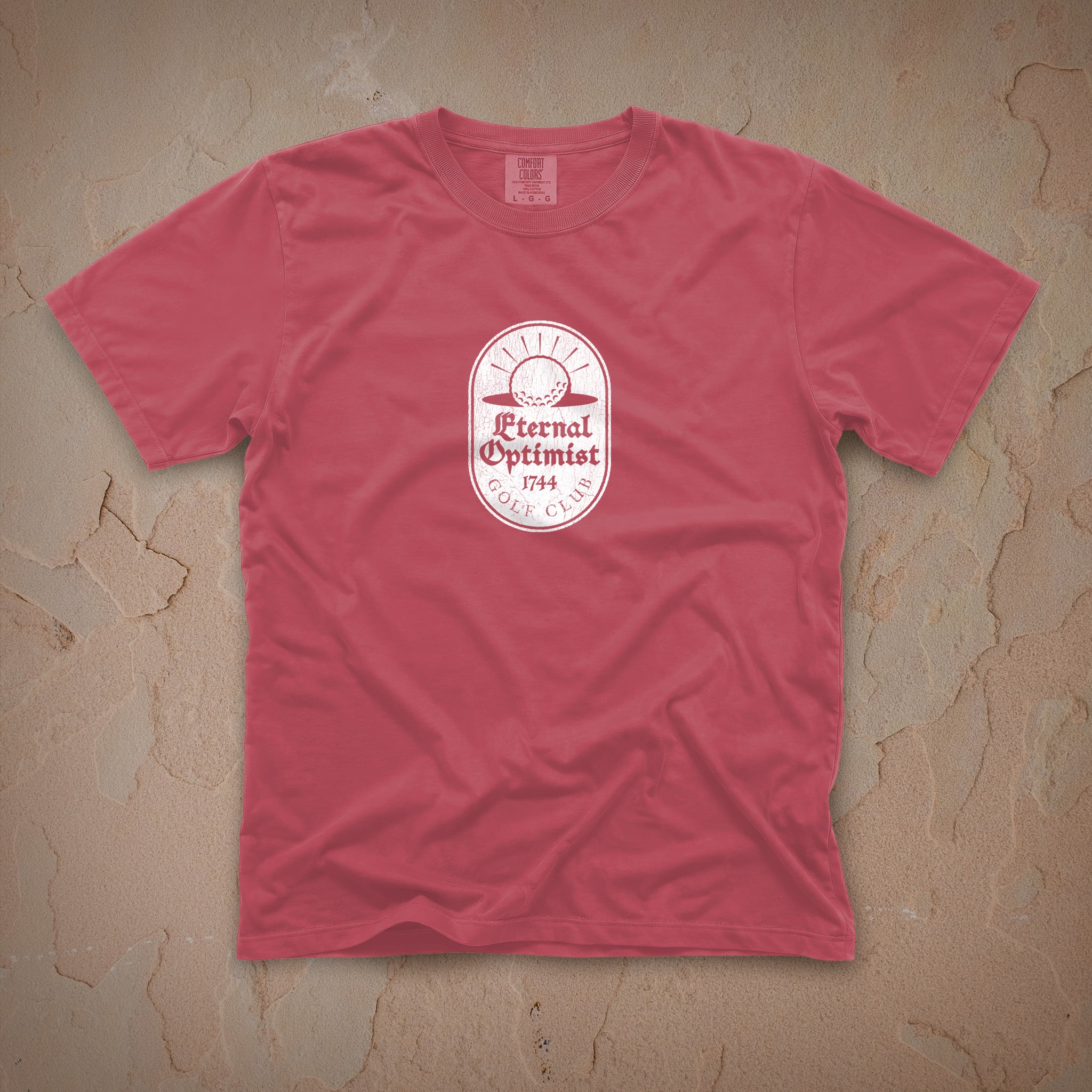 Dark pink t-shirt with a white Eternal Optimist Golf Club logo on a beige textured background.
